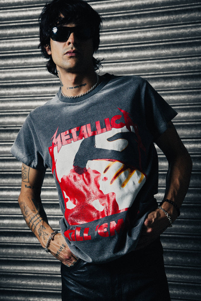 A man wearing a faded dark gray Metallica 'Kill 'Em All' graphic t-shirt, black sunglasses, and bracelets, standing in front of a corrugated metal shutter.