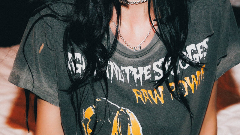 A woman with long black hair wearing a faded dark gray graphic t-shirt with yellow and white text, sitting in a dimly lit room.