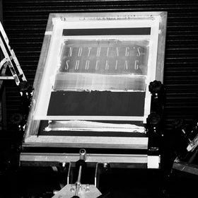 A black and white photograph showing a vintage screen printing press in use, with a wooden frame holding a printed screen and a metal press mechanism below, set against a dark corrugated metal background.