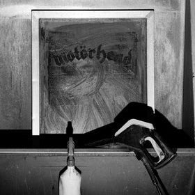 A black and white photograph showing a framed poster or print leaning against a wall on a shelf, with a spray bottle and a paintbrush nearby. The poster has the word 'motorhead' in gothic lettering.