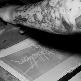 A close-up black and white photo of a tattooed arm pressing a screen to transfer a graphic onto a sheet of film or paper.