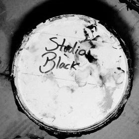 A black and white close-up of a round, worn canvas or paper disc with the words 'Studio Black' written in script across the center, surrounded by a dark rim on a textured surface.
