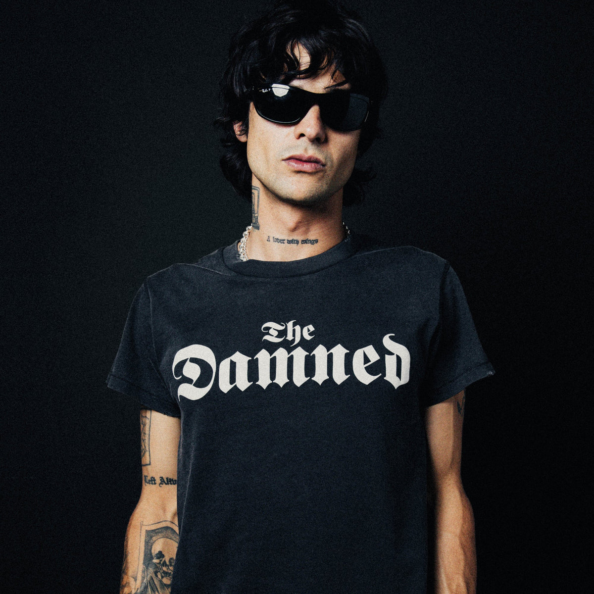 A man wearing a black graphic t-shirt with 'The Damned' printed in white gothic lettering, set against a solid black background.