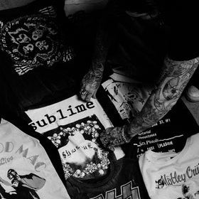A person with tattooed arms arranges a collection of black and white graphic band t-shirts on the floor. Visible shirts include Sublime, Slipknot, Korn, and Metallica.