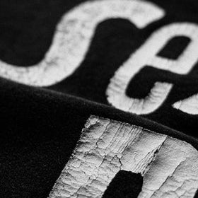 Close-up of a black fabric item with large distressed white lettering, showing part of the letters 'Se' and 'p'.