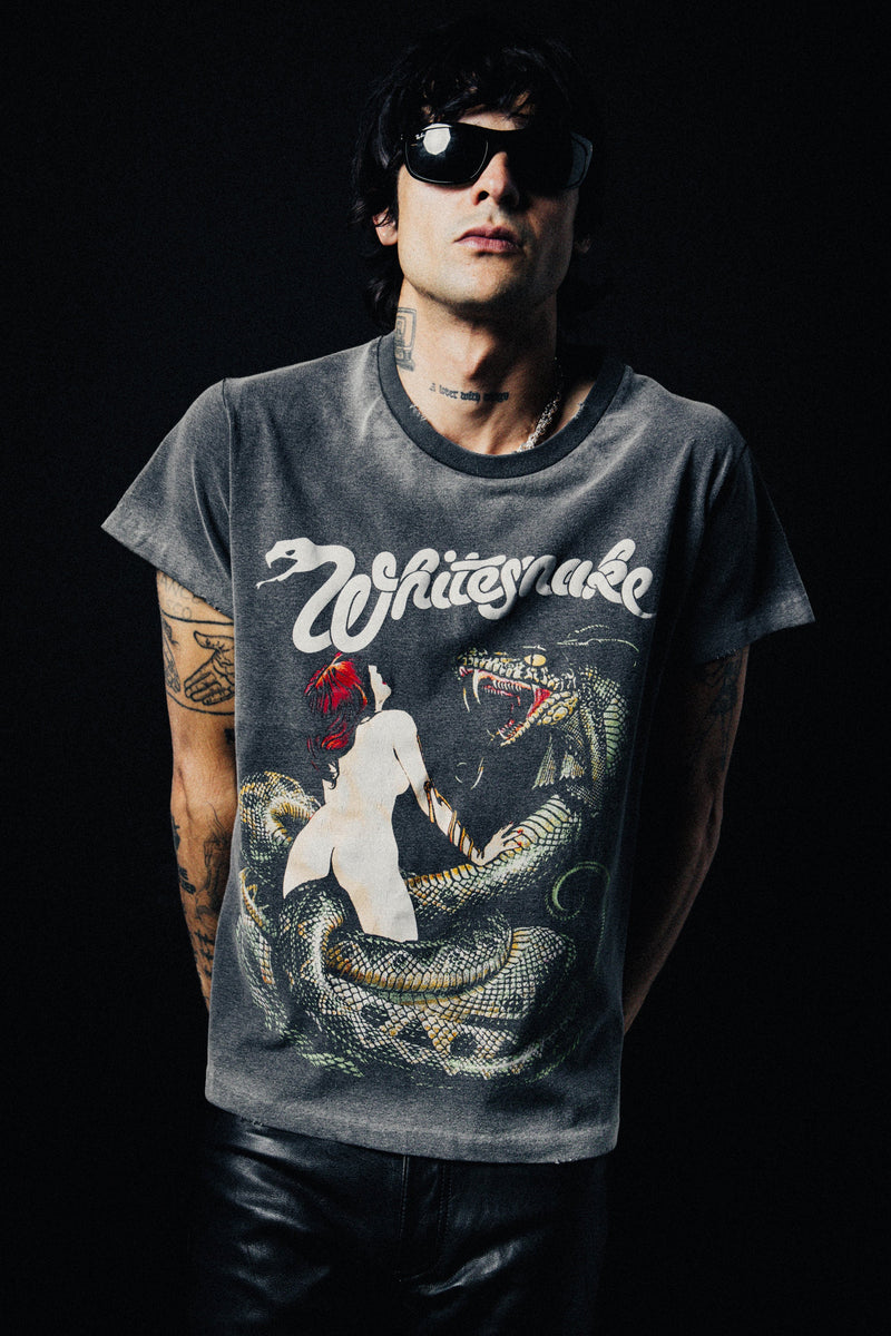 A man wearing a faded dark gray graphic t-shirt with a 'White Snake' design, styled with black leather pants and sunglasses against a black background.