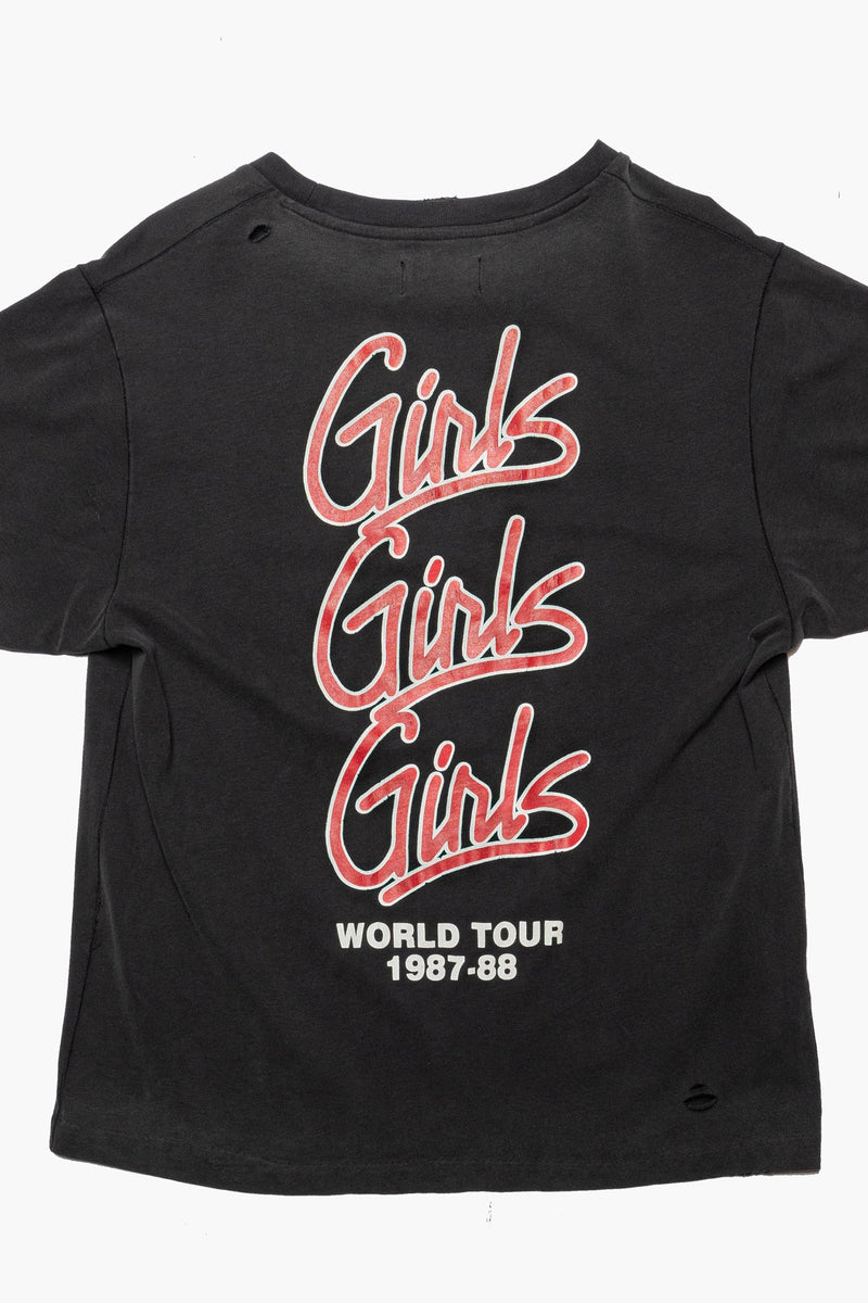 A black short-sleeve graphic t-shirt featuring the word 'Girls' repeated vertically in a pink outline script, with 'WORLD TOUR 1987-09' printed below in white, shown against a plain white background.